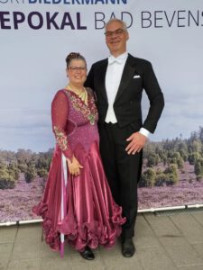 Tanzpaar Tina und Thorsten Rönnicke vor dem Plakat der Heidepokal-Turniere Bad Bevensen. Sie trägt ein beerenfarbenes, langes Kleid, er trägt einen schwarzen Frack